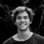 A black and white portrait of a young man smiling outdoors with natural background.