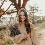 Smiling woman in cozy sweater holding a bouquet of dry flowers on a sunny outdoor path.
